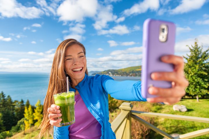 Kuvassa nuori nainen ottaa itsestään selfietä terveyspirtelön kanssa kauniissa rantamaisemassa.