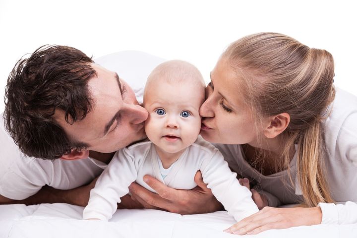 Kuvassa on perhe, johon kuuluu isä, äiti ja vauva. Vanhemmat suukottavat lasta poskille, lapsi katsoo suoraan kameraan.