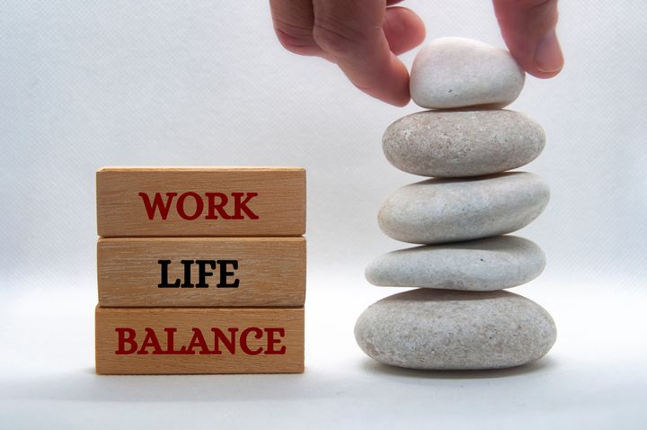 Kolme puupalikkaa, joissa tekstit "work life balance", vieressä pino pikkukiviä, käsi asettelee ylimmäistä kiveä.