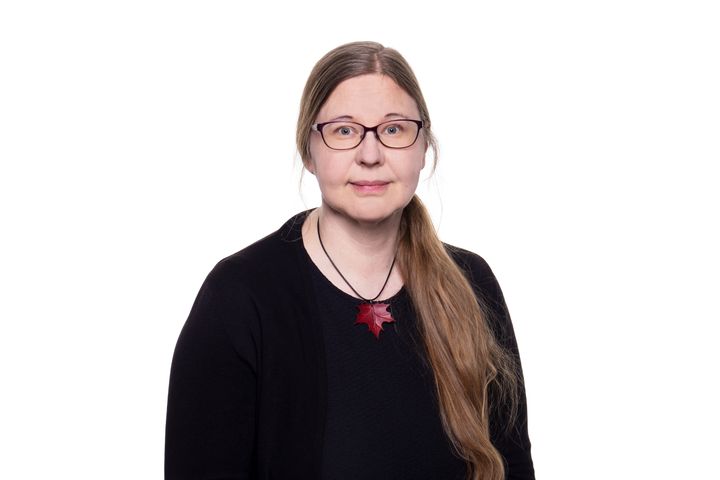 Aikuisella naisella on pitkät vaaleat hiukset, mustat vaatteet ja silmälasit. Hänellä on punainen kaulakoru. Tausta on valkoinen.