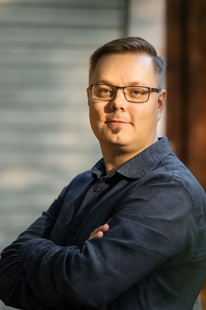 Mies seisoo, hymyilee ja katsoo kameraan. Hänellä on silmälasit ja tummat vaatteet.