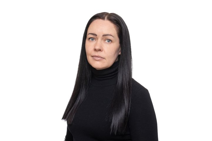 Nainen, mustat suorat hiukset ja musta poolopaita. Tausta valkoinen. Katsoo kameraan.