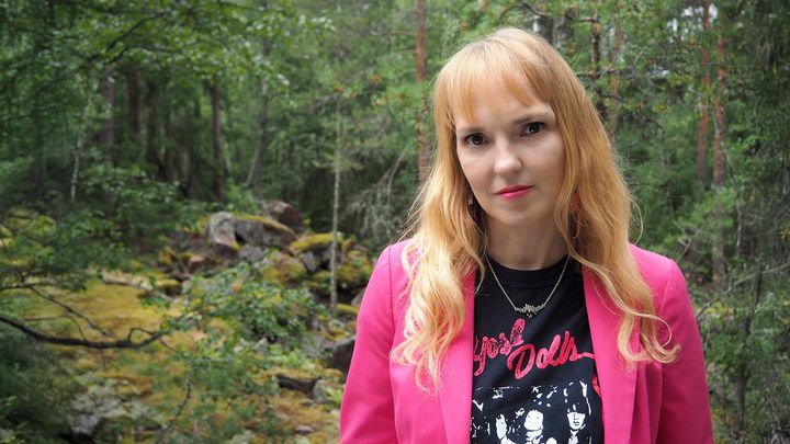 Henkilö katsoo kameraan. Taustalla metsää. Henkilöllä on pinkki takki päällä ja hiukset auki.