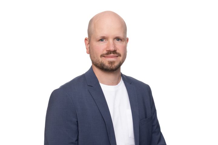 Henkilö puvuntakki päällä valkoista taustaa vasten. Hän katsoo kameraan.