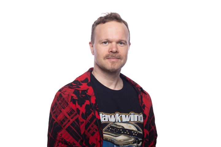 Mies katsoo kameraan. Valkoinen tausta. Päällä on musta printtipaita, jonka päällä on punamusta pitkähihainen paita.