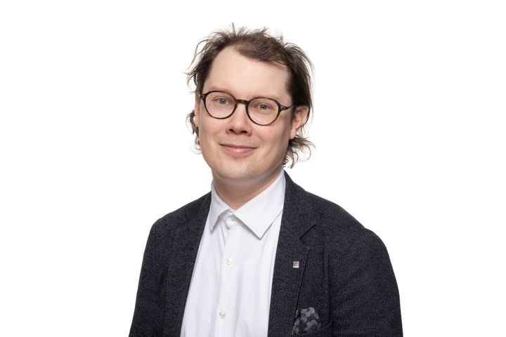 Henkilö hymyilee kameralle studiokuvassa. Päässä silmälasit ja päällä tummanharmaa puvuntakki.