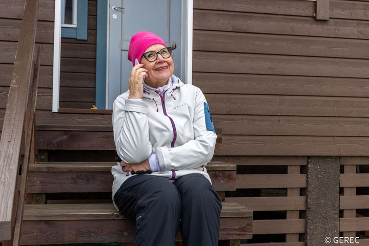 Vanhempi rouva istuu ulkoportailla ja puhuu puhelimeen. Hänellä on päällään ulkoiluvaatteet ja pinkin värinen pipo.