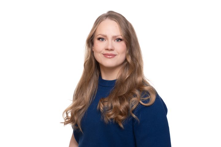 Henkilö studiokuvassa. Pitkät vaaleanruskeat hiukset ja tummansininen paita päällä.