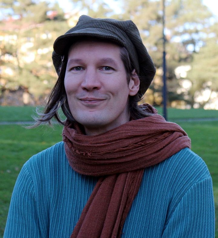 Henkilö seisoo ulkona ja katsoo hymyillen kameraan. Päällään sininen villapaita ja punaruskea villahuivi. Päässään hattu. Tummat hiukset.