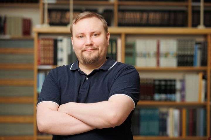 Joonas Tammela katsoo kameraan hymyillen kädet puuskassa. Hänellä on päällä tummansininen paita. Taustalla on kirjahyllyjä.