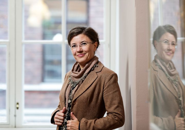 Henkilö seisoo sisällä ja katsoo hymyillen kameraan. Silmälasit ja vaaleanbeige pikkutakki. Huivi kaulassa.