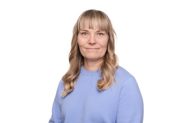 Katri katsoo kameraan hymyillen. Hänellä on pitkät vaaleat hiusket ja vaaleansininen neulepaita.