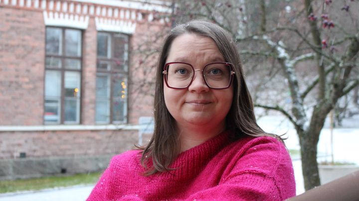 Henkilö seisoo ulkona vanhan rakennuksen edessä ja hymyilee kameralle. Taustalla näkyy ohut lumikerros. Päällä on pinkki paita ja silmälasit päässä.