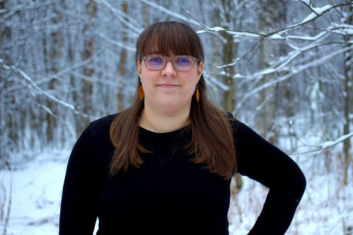 Henkilö ulkona lumisessa maisemassa. Musta paita ja silmälasit. Otsatukka.