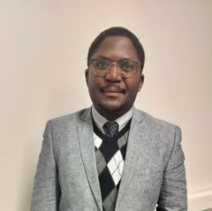 Mies seisoo ja katsoo hymyillen kameraan. Harmaa pikkutakki, liivi ja kravaatti. Silmälasit.