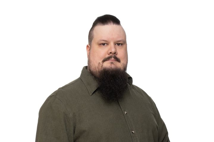 Henkilö studiokuvassa. Tummanvihreä kaulupaista ja parta.