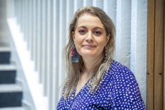 Henkilö sähkönsinisessä paidassa. Vaaleat hiukset ulottuvat olkapäiden yli. Taustalla portaikko.