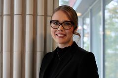 Henkilö katsoo hymyillen kameraan. Hiukset ovat kiinni, silmälasit ja musta siisti mekko.