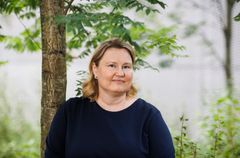Henkilö seisoo puun edessä ja hymyilee kameralle. Tummansininen yksivärinen paita.