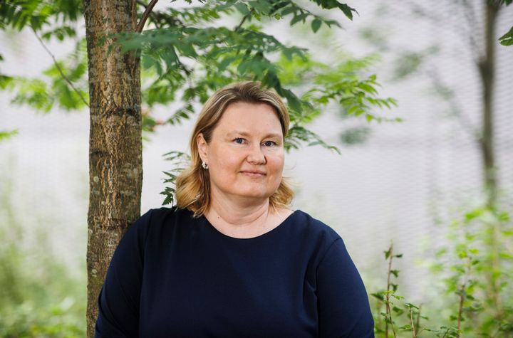 Henkilö seisoo puun edessä ja hymyilee kameralle. Tummansininen yksivärinen paita.