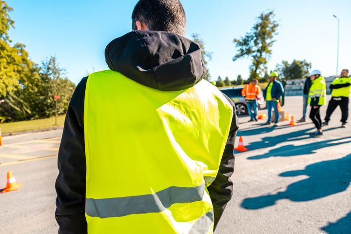 Keltainen huomioliivi päällä oleva henkilö seisoo kuvan etualalla ulkona päivän valossa selkä kameraan päin.