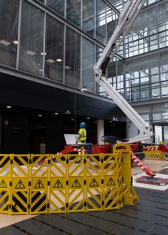 Keltaisen suoja-aidan takana on kevytrakenteinen nosturi, jonka vierellä kypärään ja huomioliiviin pukeutunut työntekijä katsoo nosturin nokkaa kohden. Taustalla näkyy toimistorakennuksen seinää.