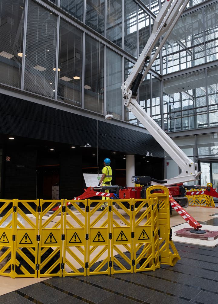 Keltaisen suoja-aidan takana on kevytrakenteinen nosturi, jonka vierellä kypärään ja huomioliiviin pukeutunut työntekijä katsoo nosturin nokkaa kohden. Taustalla näkyy toimistorakennuksen seinää.