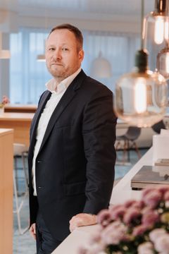 Antti Kumpulaisen puolivartalokuva. Hän seisoo sivuttain kameraan händen, katse on kameraan. Hän nojaa vasenta kättän pöytätasoon. Taustalla vaaleaa toimistotilaa.