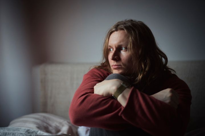 Henkilö istuu sängyllä, katselee sivulle ja halaa polviaan.