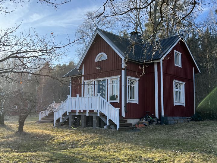 Punainen puutalo, jossa on valkoiset ikkunankarmit ja katto, seisoo nurmikolla aurinkoisena päivänä. Taustalla näkyy metsää.