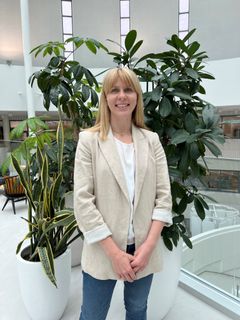 Nainen seisoo rakennuksen sisällä viherkasvien edessä vaaleassa bleiserissä ja farkuissa.