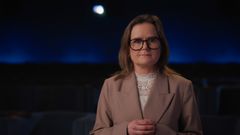 En tiedä, kuka henkilö on kuvassa. Kuvassa on henkilö, joka seisoo pimeässä huoneessa ja katsoo suoraan kameraan. Hänellä on päällään beige bleiseri ja silmälasit. Taustalla on sinistä valoa.