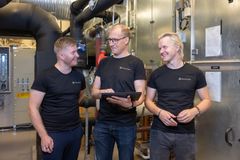 Kolme henkilöä keskustelee rakennusautomaatioon liittyvässä teknisessä tilassa. Heillä on yllään mustat t-paidat, ja yksi henkilöistä pitää tablettia käsissään.
