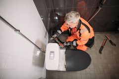 Henkilö korjaa vessanpönttöä kylpyhuoneessa oransseissa työhousuissa ja mustassa takissa.