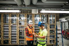 Kaksi rakennusalan työntekijää keskustelevat teknisessä tilassa, taustalla talotekniset laitteet.