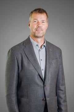 Mikael Forsblom seisoo harmaassa puvussa ja paidassa neutraalilla taustalla.