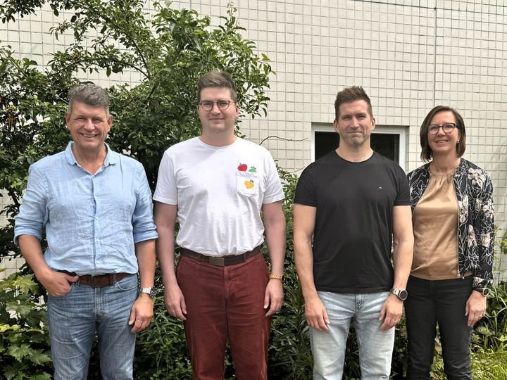 Neljä ihmistä seisoo ulkona vierekkäin, taustalla vihreitä kasveja ja vaalea seinä.