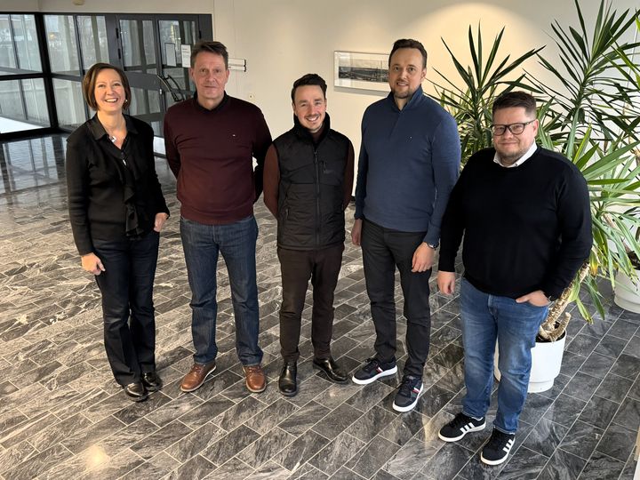 Viisi henkilöä seisoo hymyillen sisätiloissa kasvien vieressä.