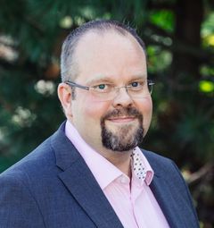 Hymyilevä mies pukeutuneena pukuun ja silmälasit, taustalla vihreitä puita.