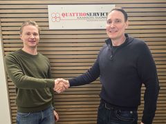 Kaksi miestä kättelee puupaneeliseinän edessä toimistossa. Taustalla on Quattroservices Kaakkois-Suomi Oy:n logo.