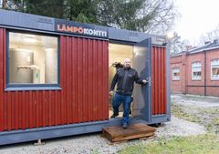 Mies seisoo punaisessa lämpökontissa, joka on sijoitettu ulkoalueelle tiilirakennuksen lähelle.