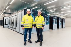 Kaksi henkilöä suojavaatteissa seisoo datakeskuksessa LUMI-supertietokoneen edessä.