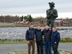 Neljä miestä seisoo Kukkolankosken rannalla Lauttamies-patsaan edessä.