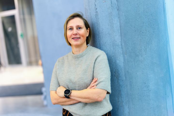 Satu Palviainen nojaa hymyillen siniseen seinään, kädet ristissä rinnalla.