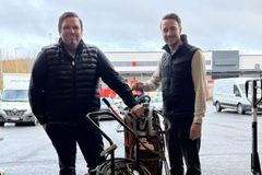 Kaksi miestä seisoo ulkona parkkipaikalla hitsauslaitteiston vierellä, taustalla pakettiautoja ja liikerakennus.