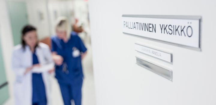 Ovessa kyltissä teksti Palliatiivinen yksikkö ja taustalla lääkäri ja hoitaja keskustelemassa.
