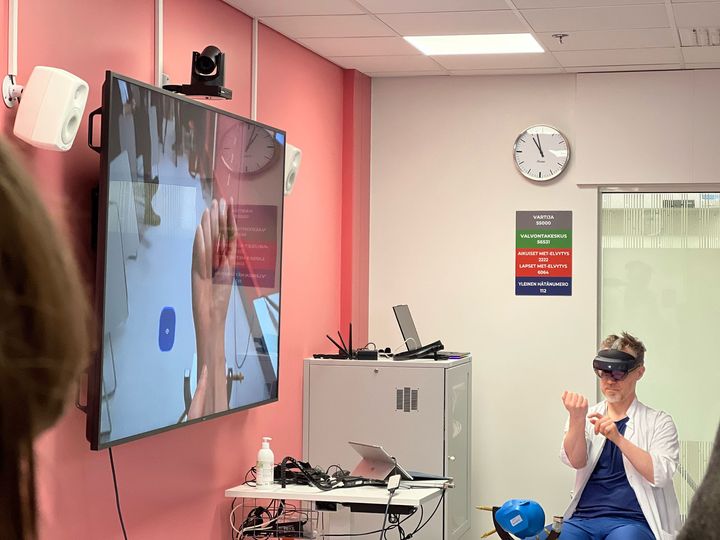 Henkilö, jolla VR-lasit istuu tuolilla. Vieressä on kuvaruutu, jossa näkyy VR-lasien näkymä.