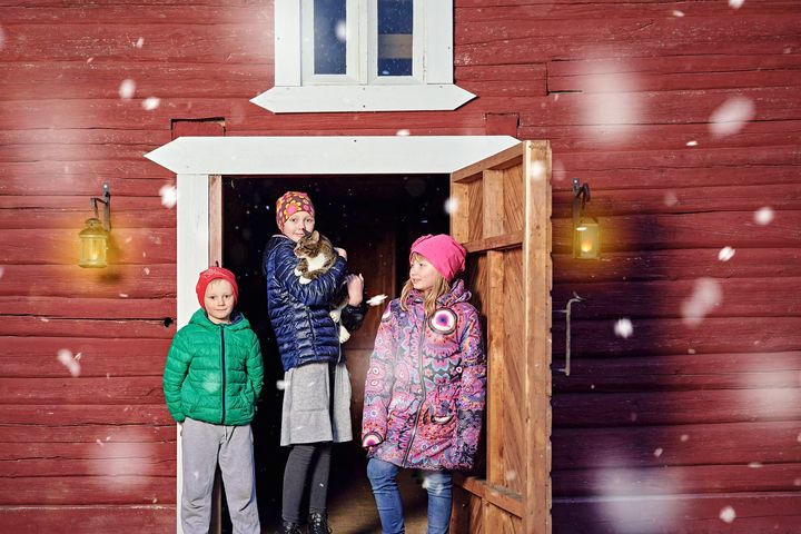 Kolme lasta seisomassa punaisen vanhemman puurakennuksen ovella ja yhdellä lapsista on kissa sylissä.