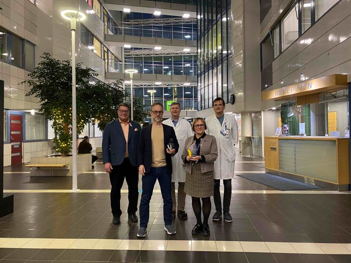 Viisi henkilöä seisoo lasiaulassa. Kahdella on palkinto kädessä.