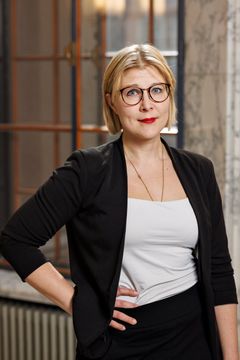 Niina Malm seisoo sisätiloissa mustassa jakussa ja silmälaseissa.